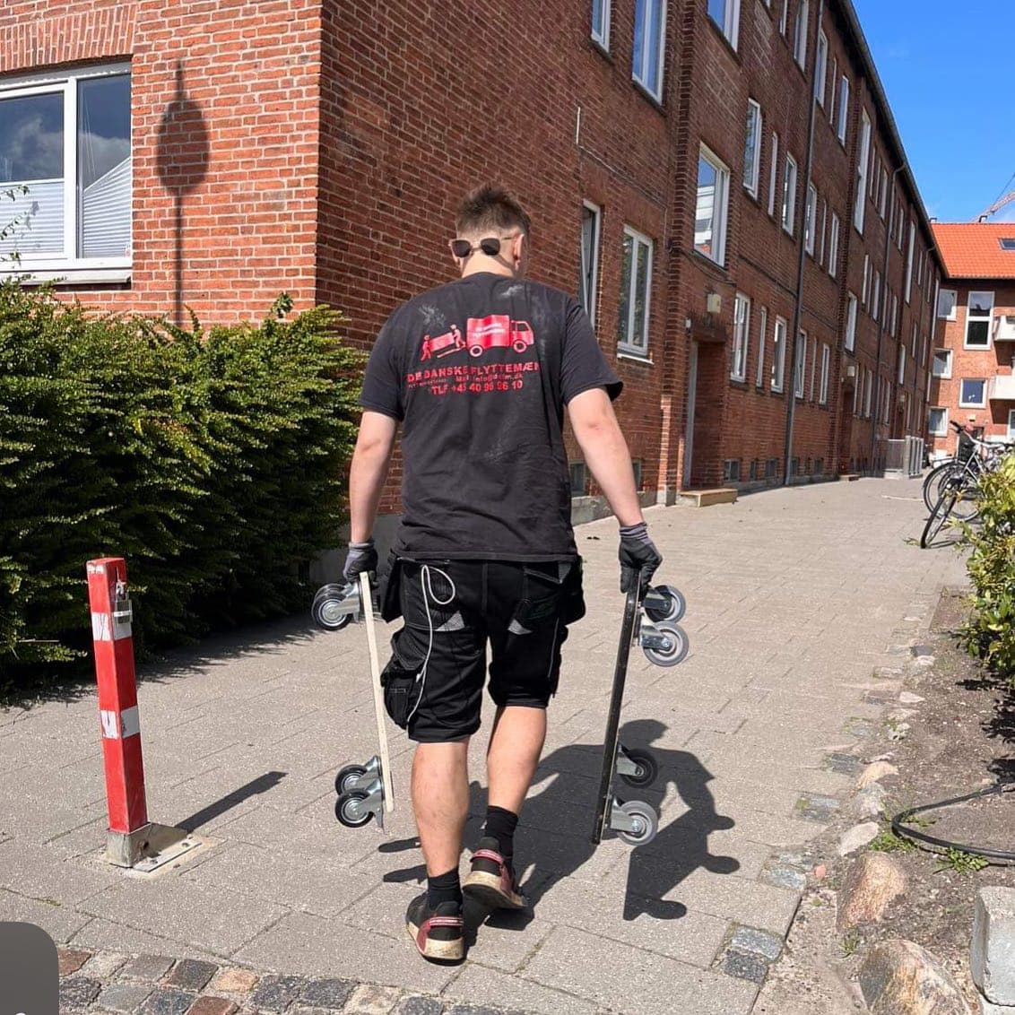 Flyttemand går med to sækkevogne på fortovet foran et rødt murstensbyggeri