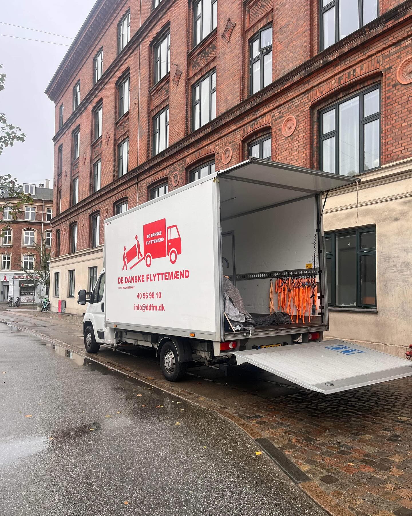 Den hvide flyttebil med åben lift holder foran en stor murstensbygning.
